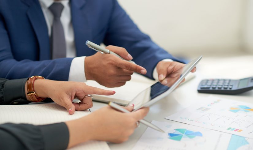 unrecognizable-business-people-sitting-at-meeting-with-charts-looking-and-pointing-at-tablet